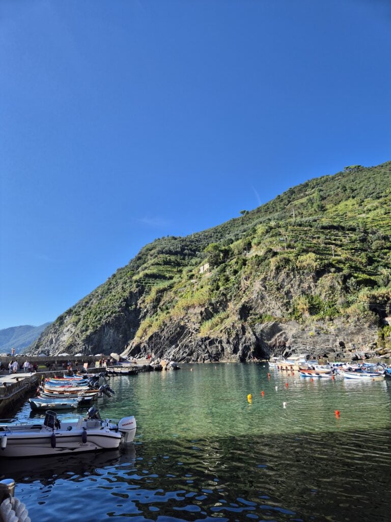 Small boats float in a calm harbor surrounded by rocky hills covered with green vegetation under a clear blue sky. People walk along the stone pier on the left side of the image.