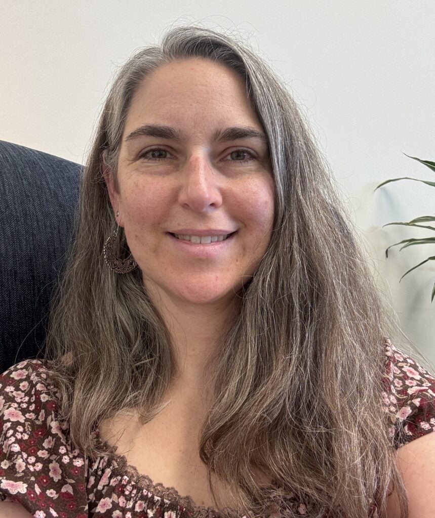 A woman with long, wavy gray hair and a floral top smiles at the camera. She is sitting indoors with a plant visible in the background.