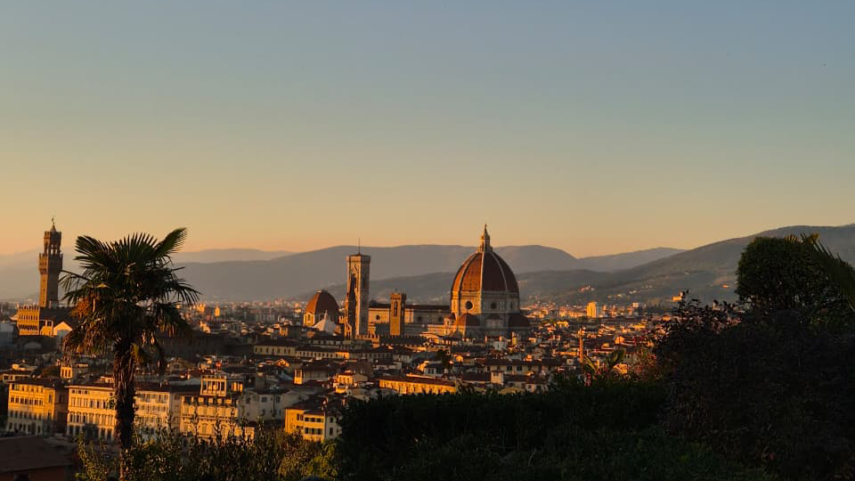 Florence skyline at sunset with the Duomo, bell tower, and city buildings visible. Mountains are in the background, and trees frame the foreground.