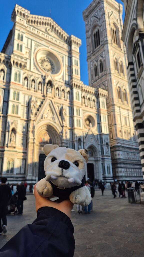 A hand holds a small plush bulldog toy in front of the ornate facade of Florence Cathedral, with people walking and the bell tower visible in the background under a clear blue sky.