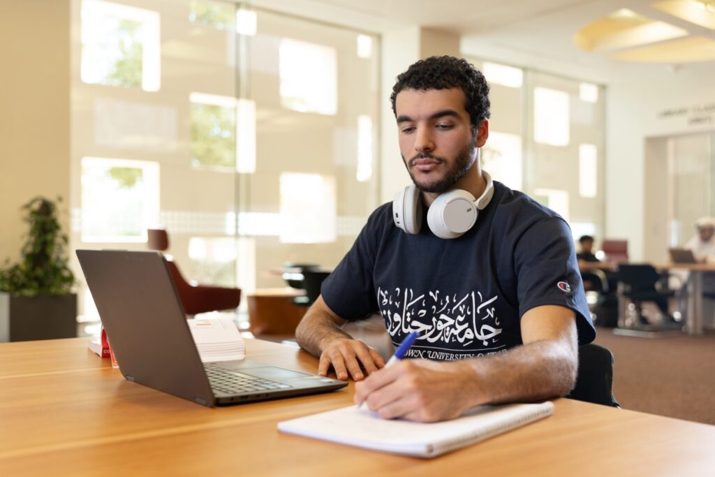 A young man wearing headphones sits at a table in a bright study area, working on a laptop and writing in a notebook. There are books and people in the background.