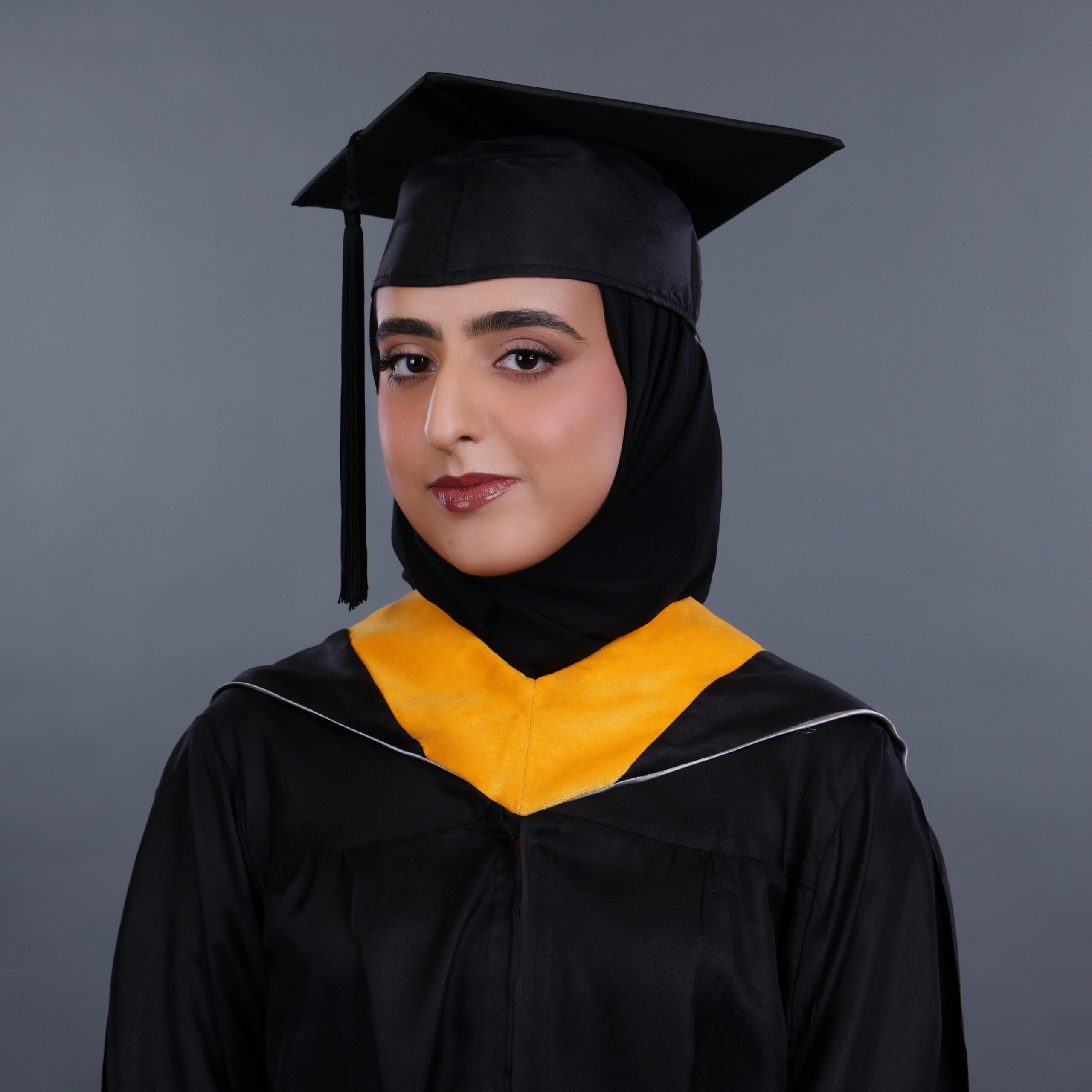 A woman wearing a black graduation gown and cap with a yellow stole smiles slightly, posing in front of a plain gray background. She is also wearing a black hijab under her cap.
