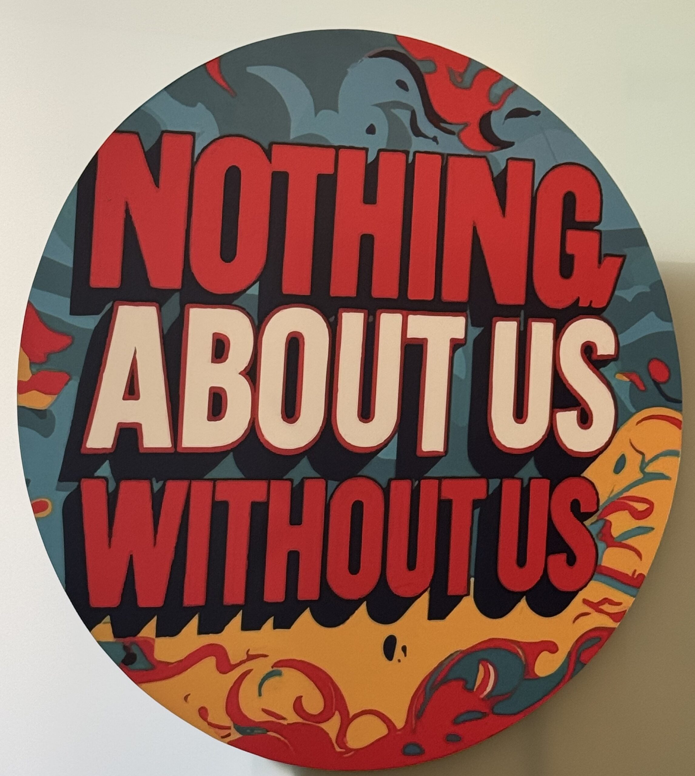 A round sign with bold red and white letters reads, NOTHING ABOUT US WITHOUT US, against a colorful background with abstract blue, yellow, and red designs.