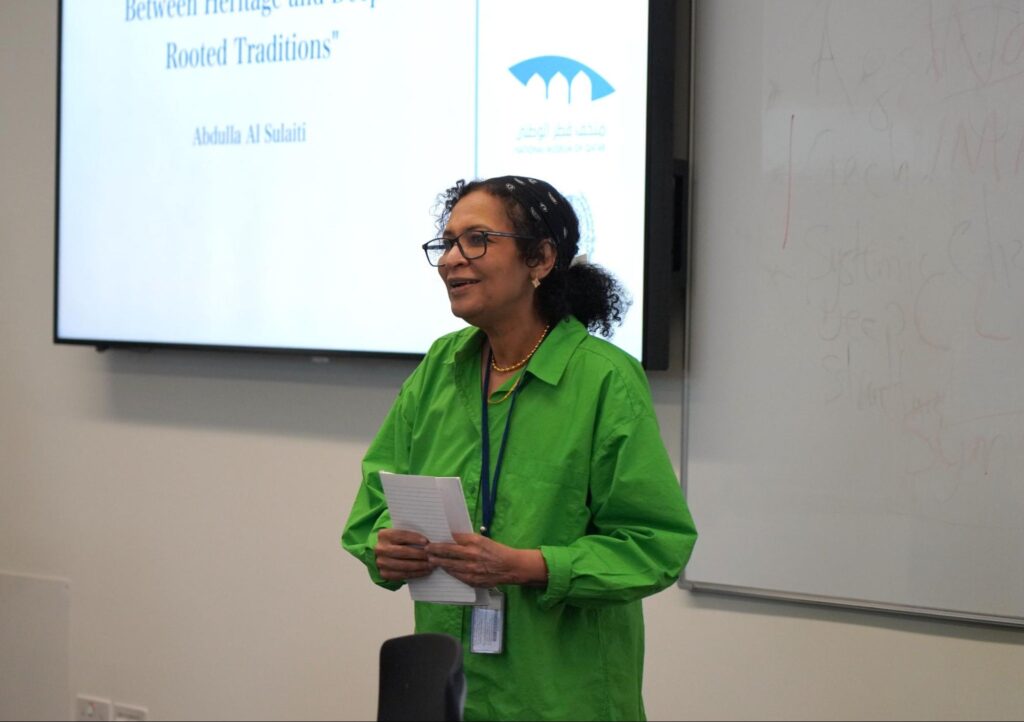 A woman in a bright green shirt holds papers and speaks in front of a screen displaying a presentation titled Between Heritage and Deep-Rooted Traditions by Abdulla Al Saiti.