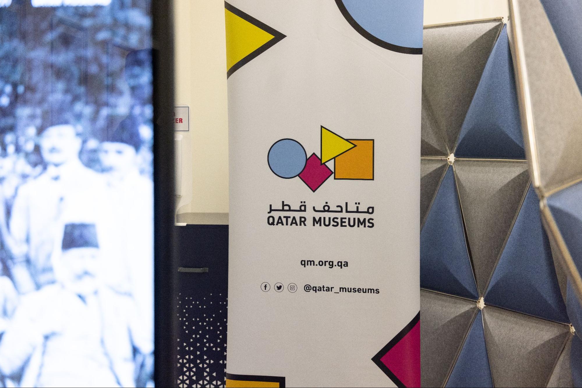 A Qatar Museums banner with geometric shapes stands beside a blurred black-and-white photo and a blue geometric-patterned wall panel.