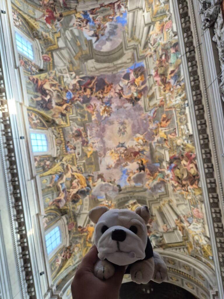 A hand holds a small plush bulldog toy in front of a grand, ornate church ceiling decorated with colorful baroque frescoes and architectural details.