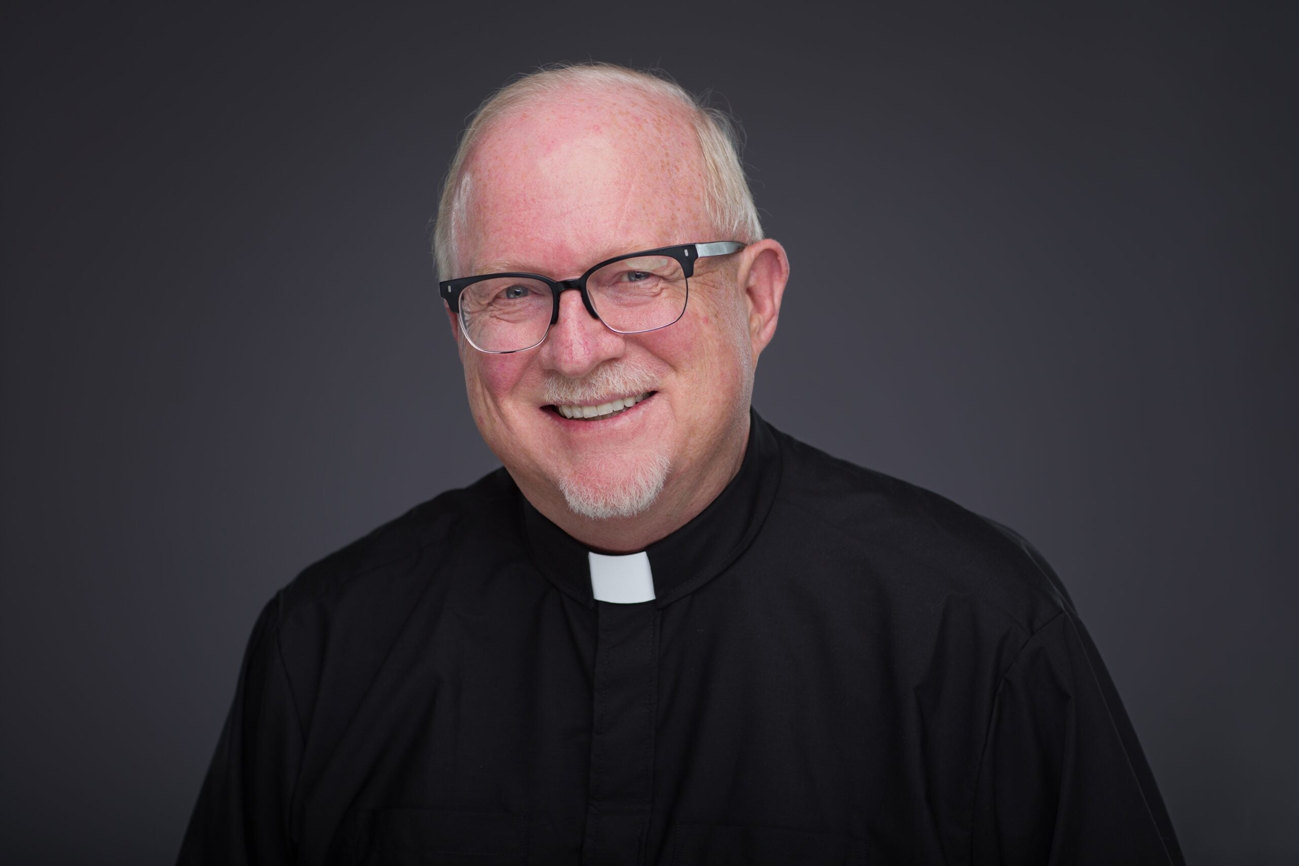 Smiling older man with glasses, wearing a black clerical shirt and white collar, posed against a plain dark gray background.