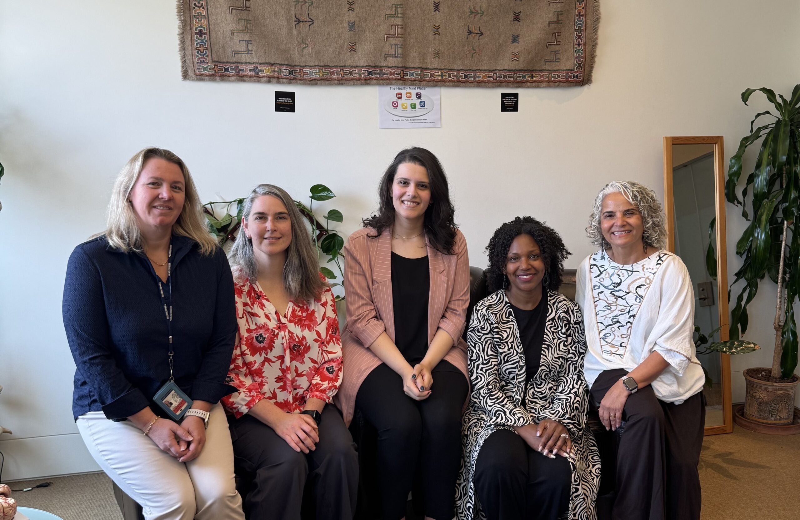 Five women sit side by side indoors, smiling at the camera. There are plants around them and a decorative rug hanging on the wall behind them.
