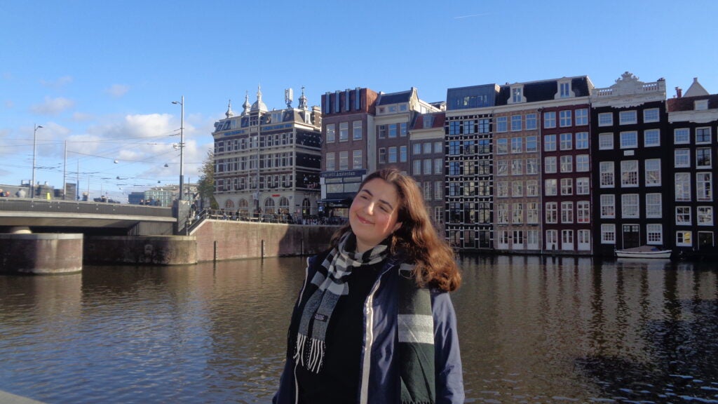 A smiling person stands by a canal in Amsterdam, with traditional Dutch buildings and a bridge in the background under a clear blue sky.