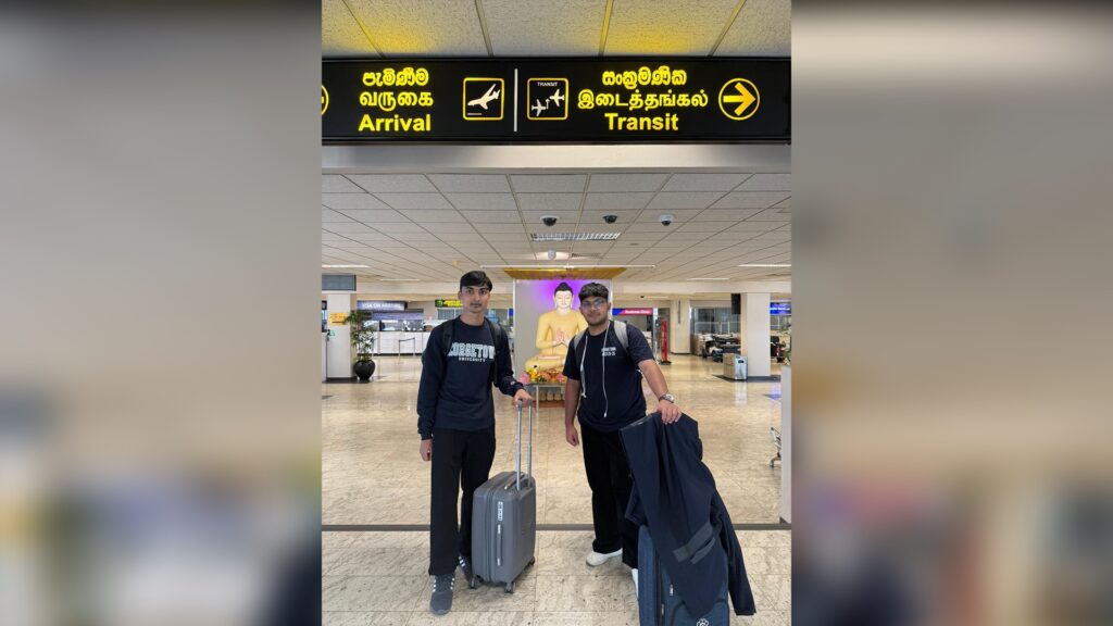 Two people stand in an airport terminal with suitcases under a yellow sign showing directions for Arrival and Transit in English and two other languages. A Buddha statue is visible in the background.