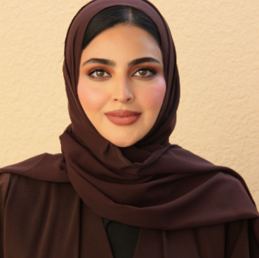 A woman wearing a dark brown hijab and matching outfit smiles softly at the camera against a light beige textured wall.