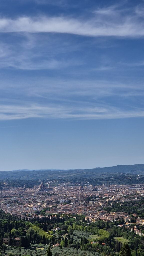 A panoramic view of a city with dense buildings, surrounded by green trees and hills under a blue sky with wispy clouds.