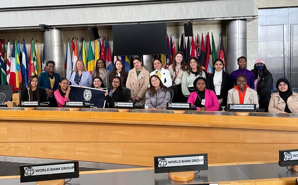 A group of diverse young women pose together in a conference room with international flags behind them and World Bank Group signs on the tables.