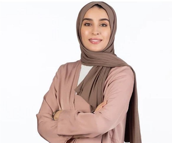 A woman wearing a light brown hijab and a pale pink jacket stands with her arms crossed, smiling at the camera against a plain white background.