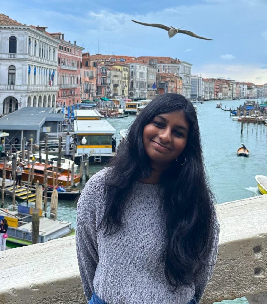 A young woman with long dark hair smiles on a bridge above a canal in Venice, Italy; colorful buildings, boats, and a flying seagull are visible in the background.