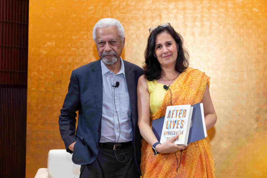 A man in a suit stands beside a woman in a yellow sari holding a book titled AFTERLIVES. Both are wearing microphones and smiling in front of a textured yellow background.