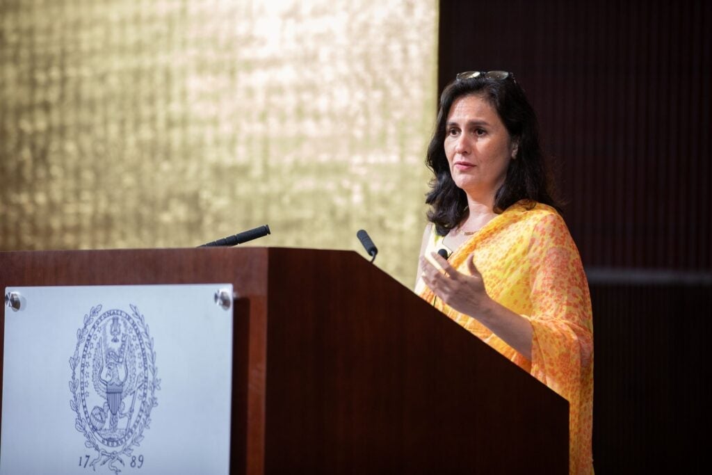 A woman in an orange saree speaks at a podium with a crest, standing in front of a gold and brown background.