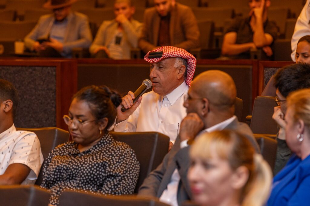 A man in a red and white keffiyeh speaks into a microphone during an event, seated among a diverse audience in an auditorium.