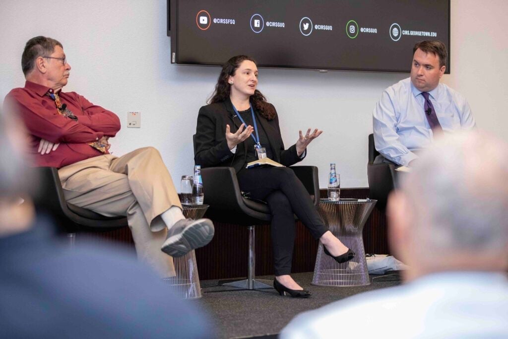 Three people sit on stage in chairs at a panel discussion. The woman in the center speaks with expressive hand gestures, while the two men on either side listen. A screen with social media icons is visible behind them.
