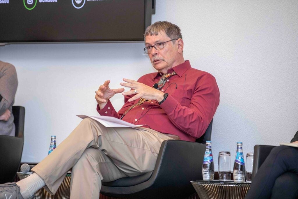 A man in a red shirt and khaki pants sits in a chair, speaking and gesturing with his hands during a panel discussion. Bottled water and papers are on nearby tables.