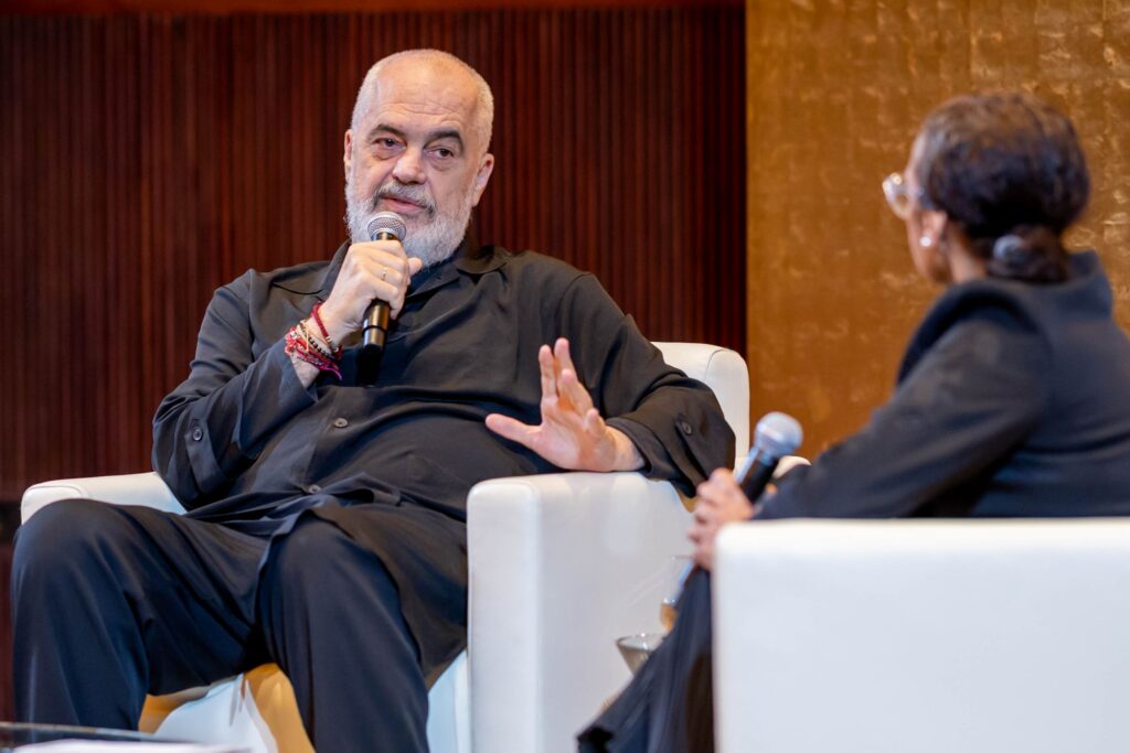 A man with a gray beard speaks into a microphone while seated on a white chair, gesturing with his hand. Another person holding a microphone sits across from him. Both are dressed in dark clothing.