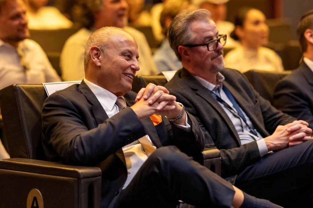 Two men in suits sit in an audience, smiling and appearing engaged. Several people are seated in the background, slightly out of focus. The setting appears to be a formal event or conference.