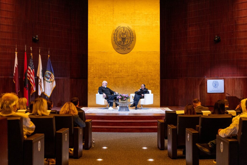 Two people sit on stage having a discussion in an auditorium with an audience watching. Several flags are displayed to the left, and a large gold emblem is on the wall behind the speakers.