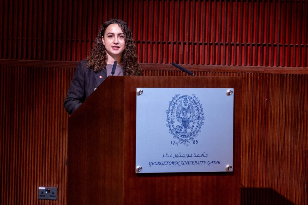 A person with curly hair speaks at a wooden podium with the Georgetown University Qatar seal and name, set against a wooden-paneled wall.