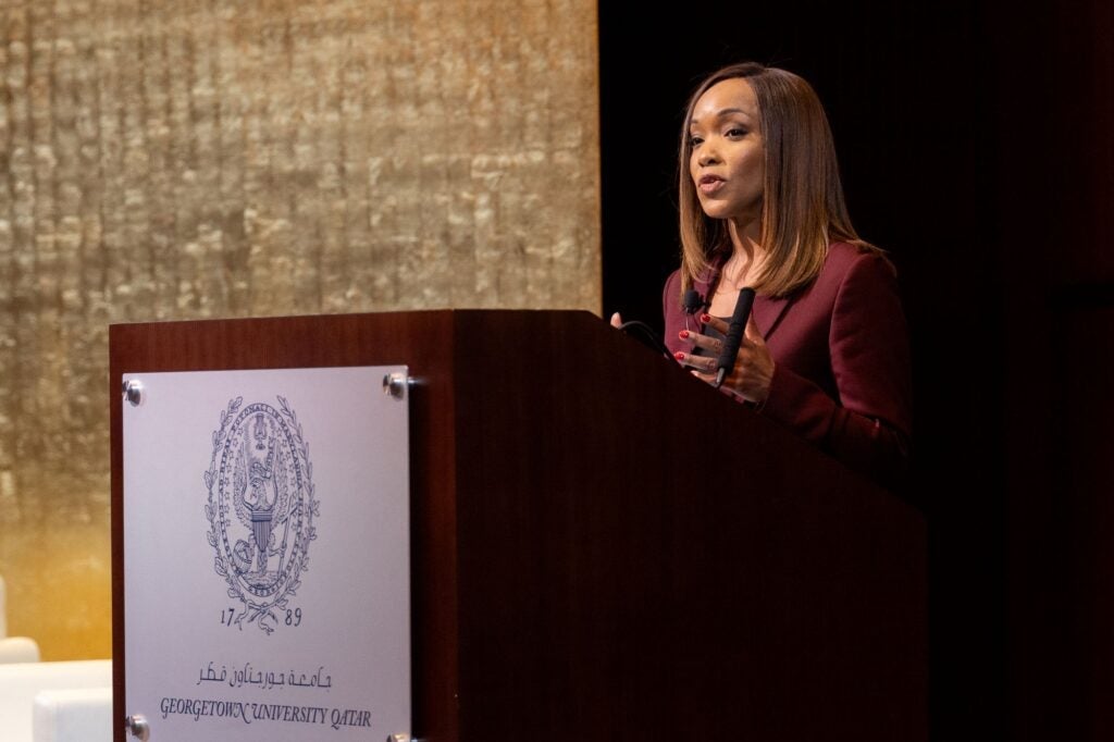 A woman in a maroon blazer speaks at a podium with the Georgetown University Qatar emblem, against a gold-toned background.