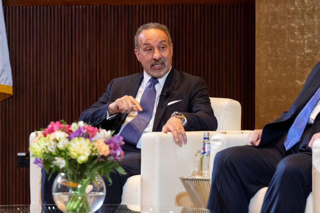 A man in a suit sits in a white armchair, gesturing while speaking. There is a bouquet of flowers and a bottle of water on the table in front of him. Another person sits nearby, partially visible.