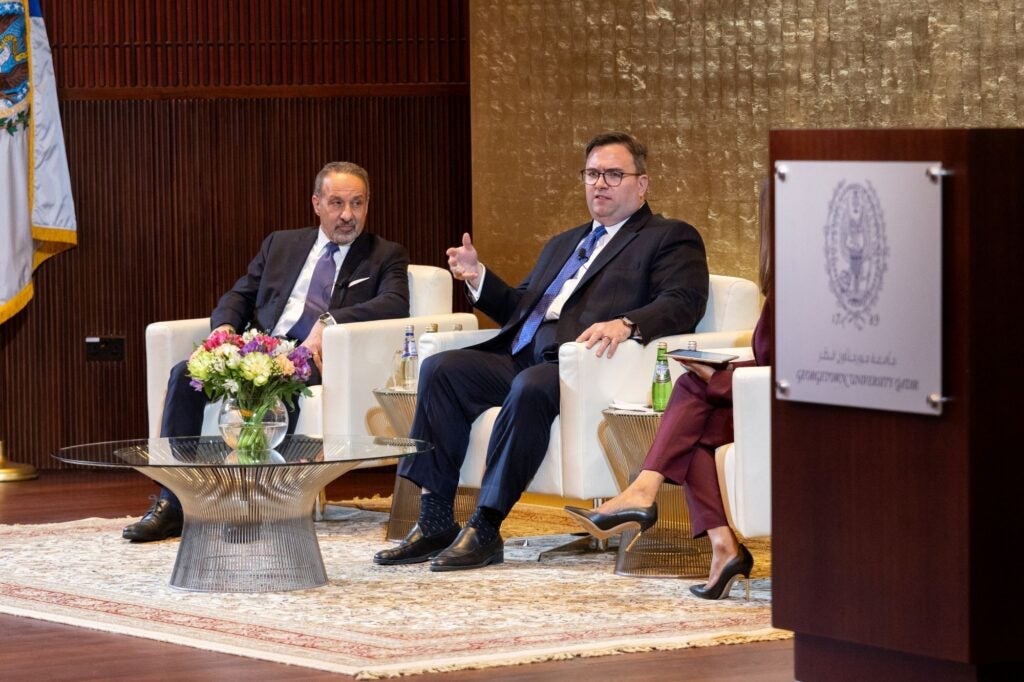 Two men in suits sit on white chairs, speaking at a panel discussion. A glass table with flowers is in front of them. A third persons legs are visible. A podium and gold-textured wall appear in the background.