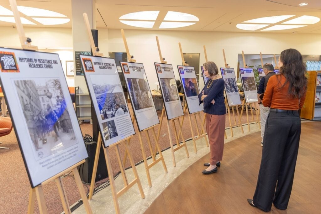 Two women view a row of display boards with photos and text on easels in a well-lit indoor exhibition space. Other people are visible further along the display.