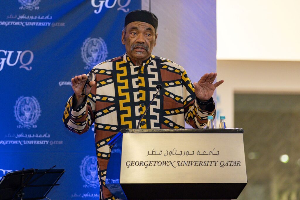 A man in a colorful patterned shirt and black hat speaks at a podium labeled Georgetown University Qatar with a blue backdrop featuring the universitys logo.