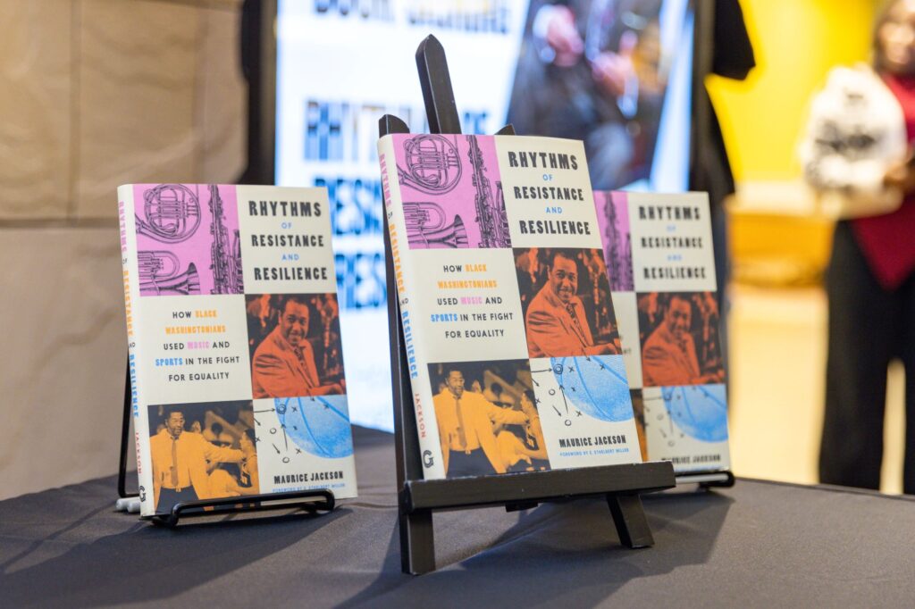 Three copies of the book Rhythms of Resistance and Resilience by Maurice Jackson are displayed upright on a table, with colorful covers featuring photos of protest and sports scenes.