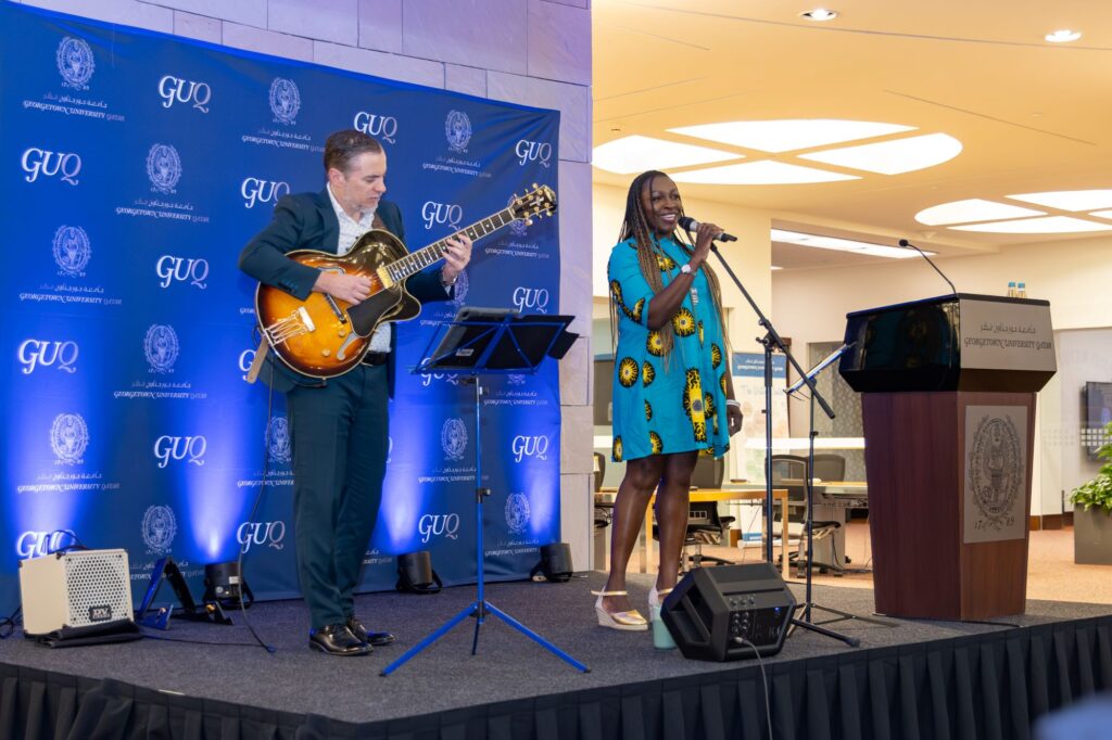 A guitarist in a blue suit plays while a woman in a blue patterned dress sings onstage at a Georgetown University event, with a blue GUQ backdrop and a podium nearby.