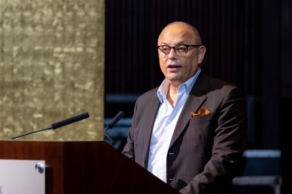 A man in a suit and glasses speaks at a podium with a microphone, standing in a formal indoor setting with a gold-colored background.