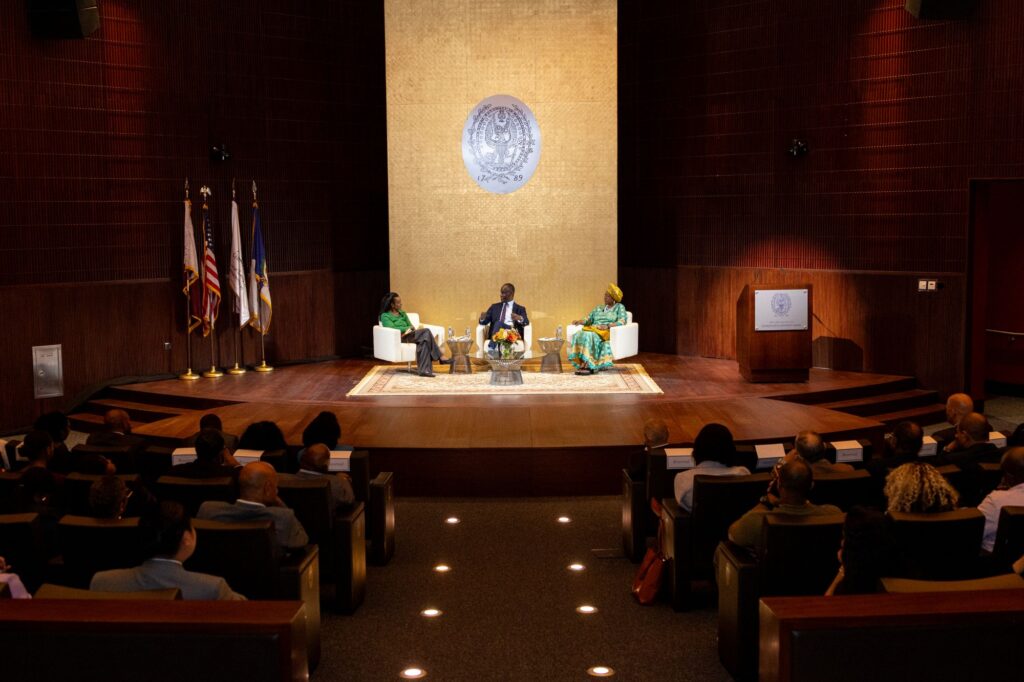 Three people sit onstage in armchairs, having a discussion in front of an audience in an auditorium. U.S. and state flags are displayed on the left, and a large gold backdrop with an emblem is behind the speakers.