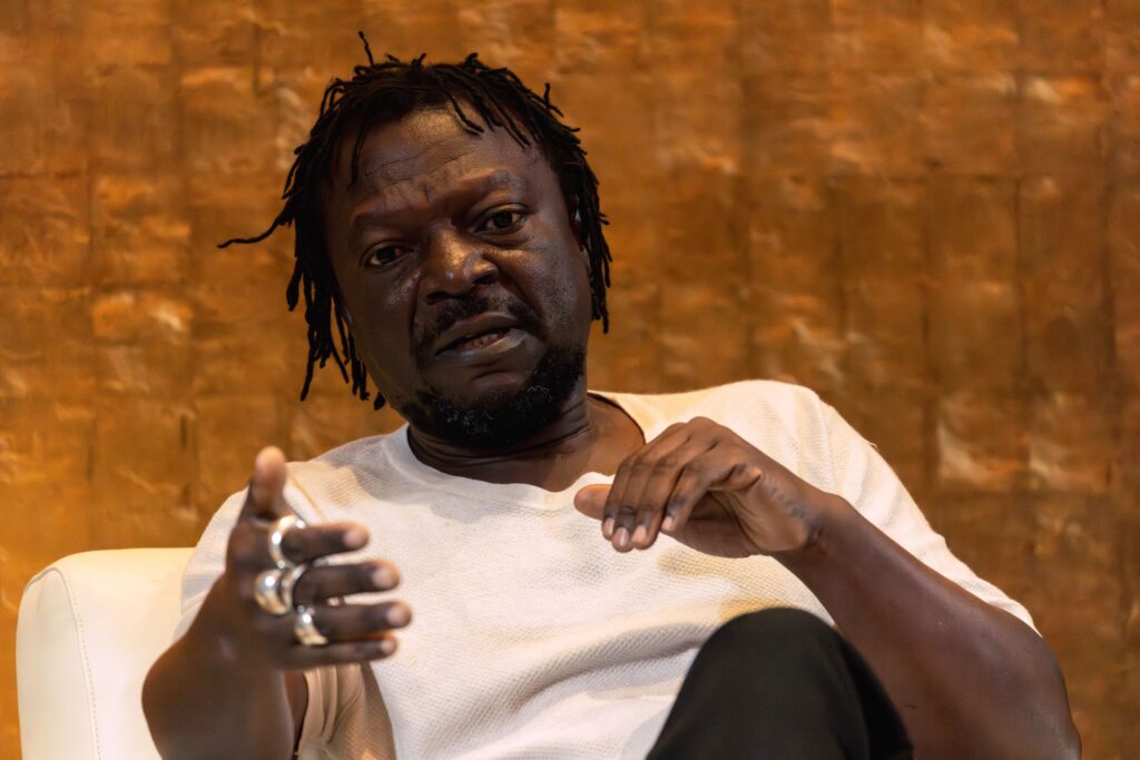 A man with short dreadlocks and facial hair sits on a white chair, wearing a light shirt and rings, gesturing with one hand as he speaks. The background is a textured brown wall.