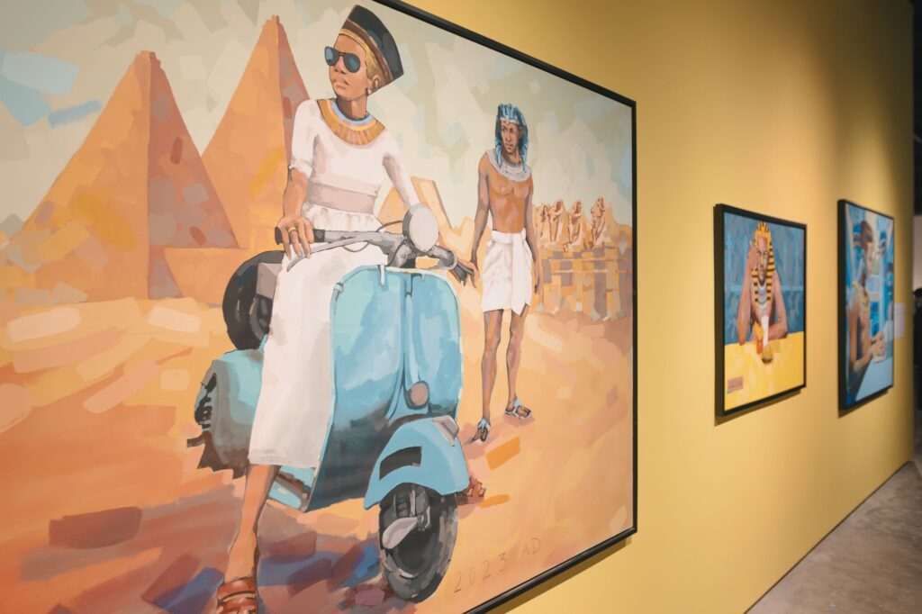 A painting of an ancient Egyptian queen in modern clothes rides a blue scooter, with pyramids and a man in traditional attire in the background. Other Egyptian-themed artworks hang on the yellow wall.