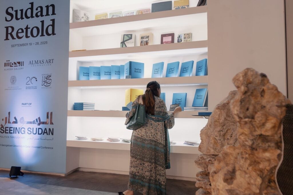 A woman in a patterned dress browses blue books on white shelves at the Sudan Retold exhibit, with informational text and logos displayed on a white wall to her left.