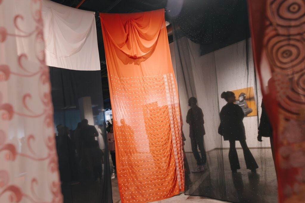 Sheer fabric panels in various colors hang in a gallery space, with silhouettes of people visible behind them and artwork displayed on the walls.