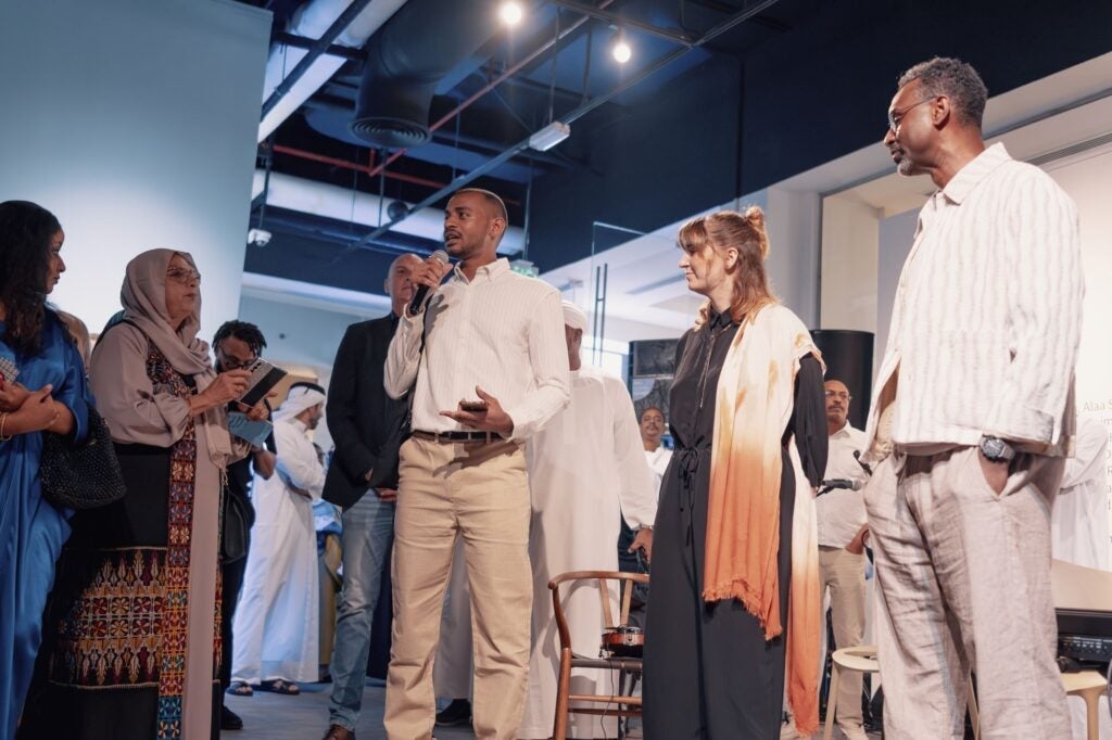 A man speaks into a microphone, surrounded by a diverse group of people in a modern indoor setting. Some listen attentively while others watch, with artwork and lights visible in the background.