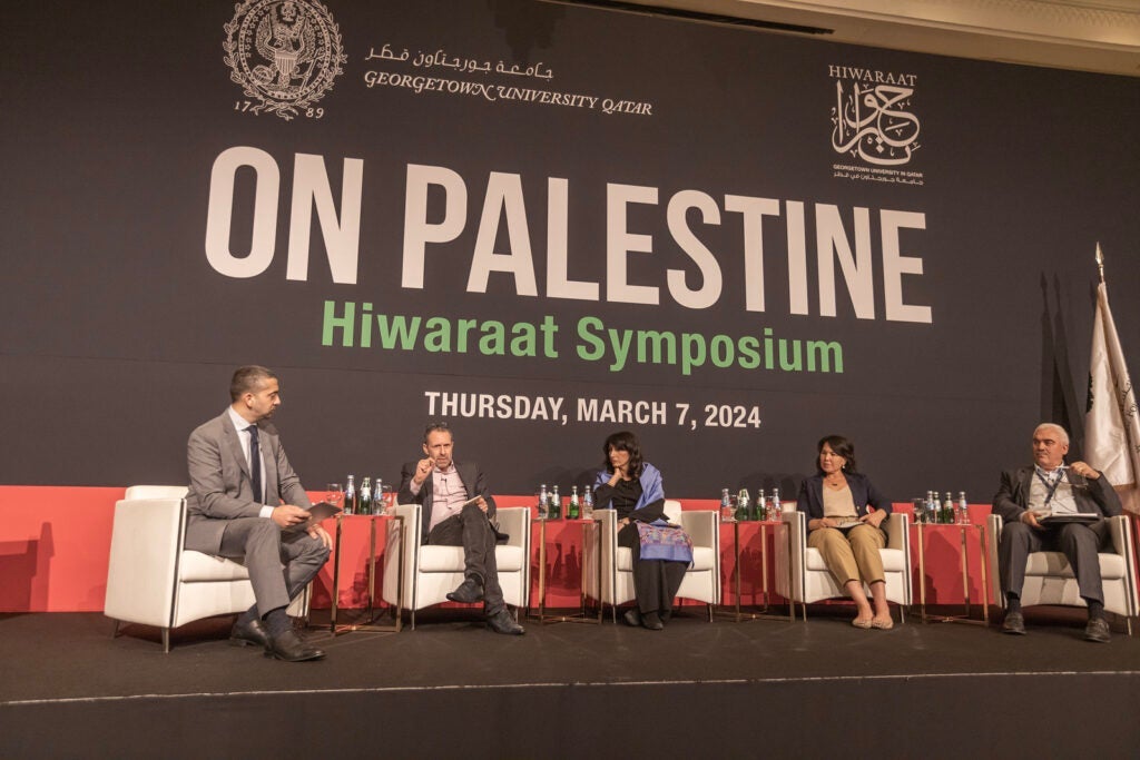 Five panelists seated on stage at the Hiwarraat Symposium, Georgetown University Qatar, discussing Palestine on March 7, 2024, with a large event banner in the background.