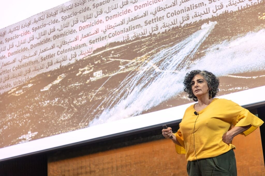A woman in a yellow top stands in front of a screen displaying words like Domicide and Settler Colonialism alongside an image of land, speaking with a microphone.