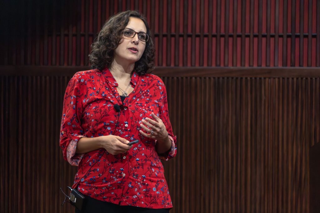 A woman wearing glasses and a red floral blouse speaks in front of a wooden panel background, holding a small device and gesturing with her hand.