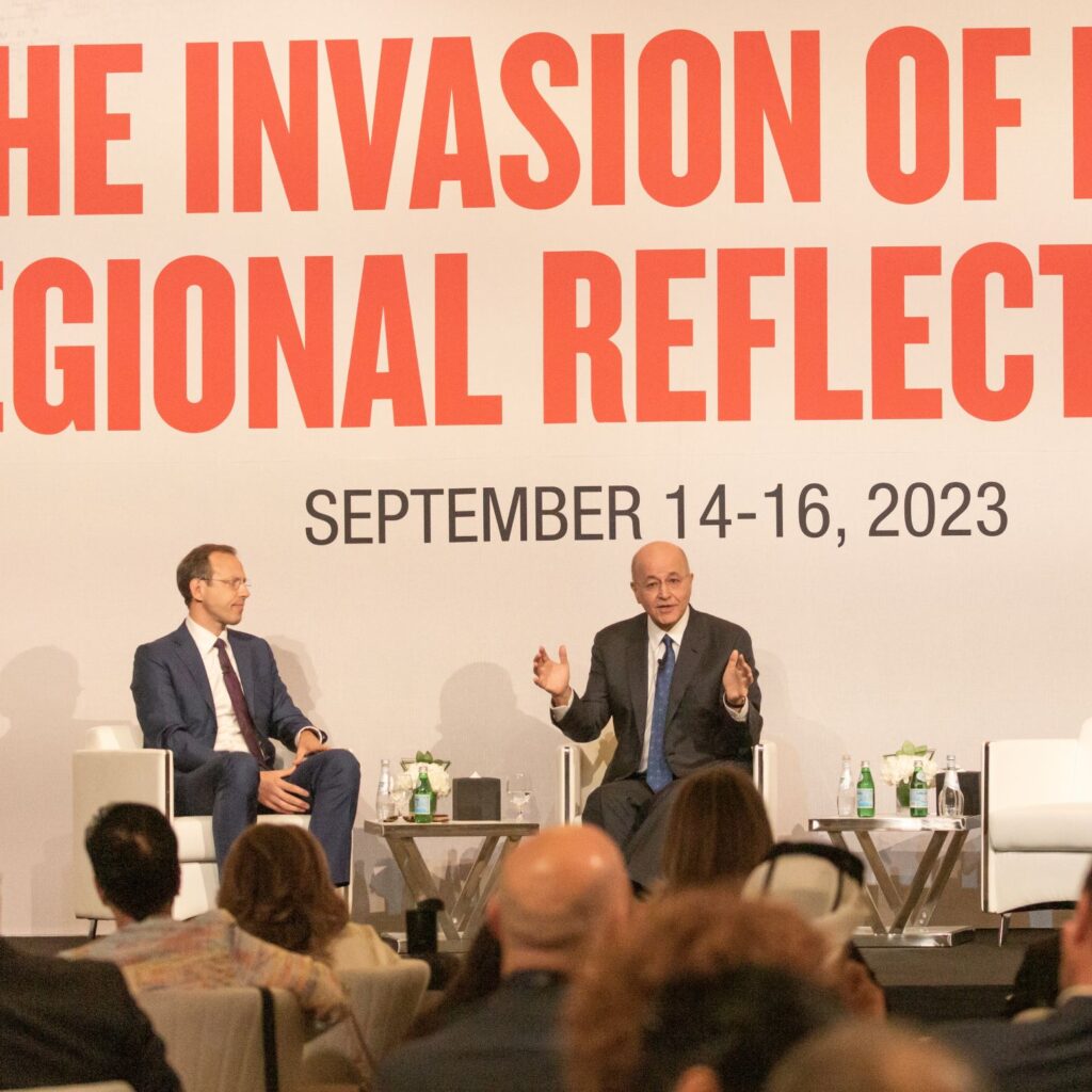Two men in suits sit on stage in front of an audience, speaking at an event. The backdrop reads, THE INVASION OF R... REGIONAL REFLECT... SEPTEMBER 14-16, 2023..