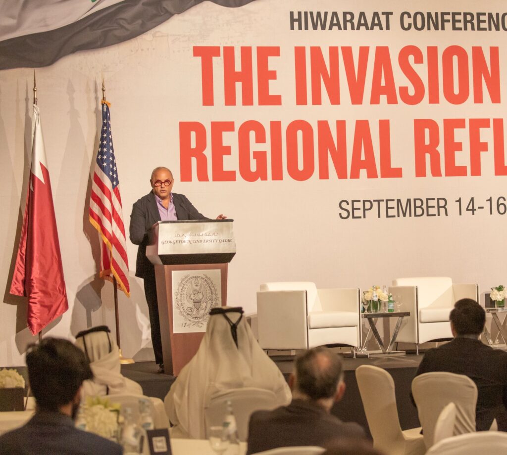 A speaker at a podium addresses an audience at a conference, with Qatari and U.S. flags nearby and a large banner reading THE INVASION & REGIONAL REF and event dates in the background.