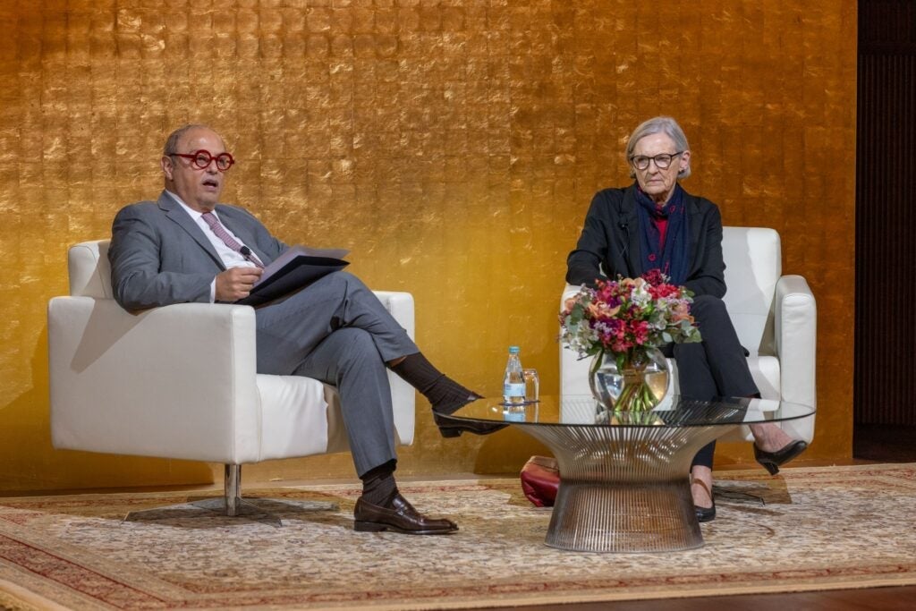 Two people sit on white chairs on a stage with a gold textured backdrop. A small table with flowers and water bottles is between them. One person holds papers; both appear engaged in conversation.