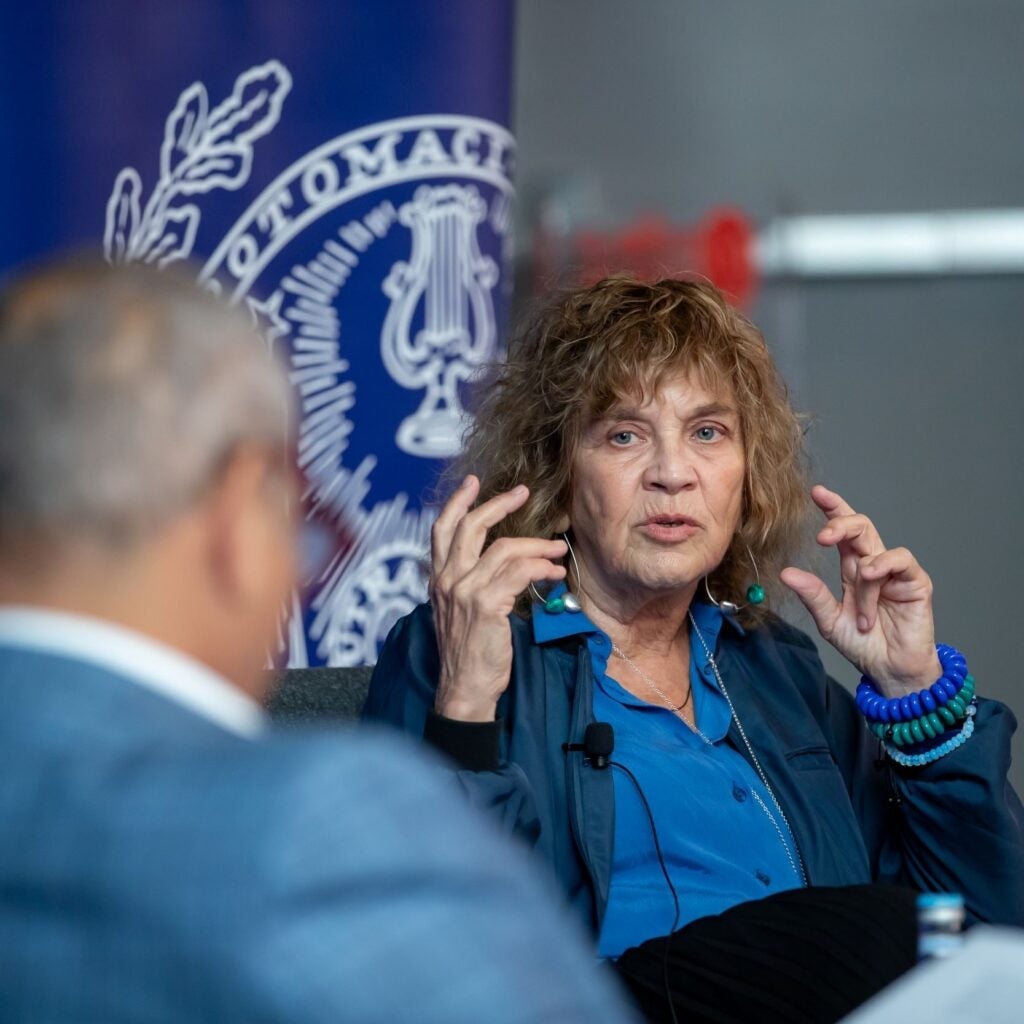 A woman with curly hair gestures while speaking during a discussion panel. She wears a blue shirt, blue bracelets, and sits in front of a blue banner with a white emblem. Another person is blurred in the foreground.