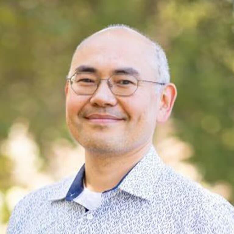 A man with glasses and a short beard smiles gently at the camera, wearing a patterned white shirt. The background is softly blurred greenery with warm light.